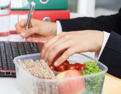 A person in business attire works on a laptop with a notebook, while eating lunch.