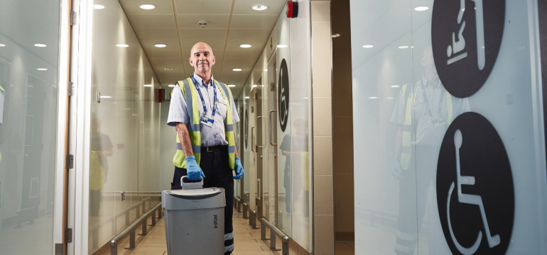 A hygiene technician carrying a sanitary waste bin through an accessible washroom corridor, representing professional washroom maintenance and inclusivity.