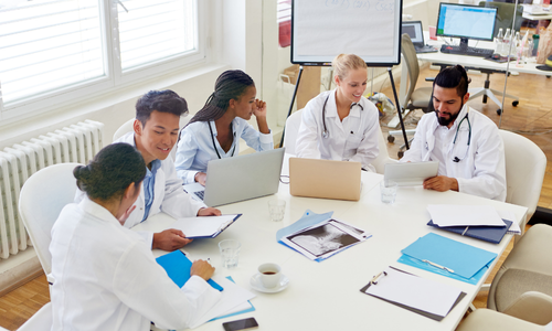 Five medical professionals in white coats sit around a table with laptops, documents, and x-rays, engaged in discussion in a bright office.