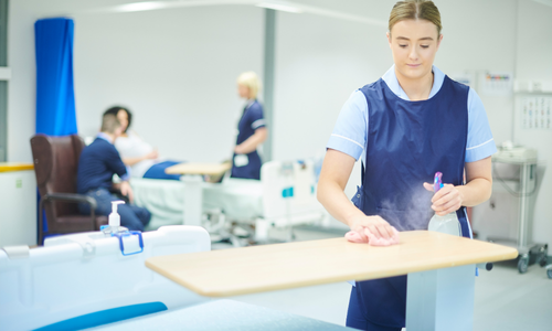 A healthcare worker wearing a blue uniform and gloves disinfects a hospital bedside table using a spray bottle and cloth. In the background, other medical staff and a patient are visible in a hospital ward setting, emphasising cleanliness and infection control practices.
