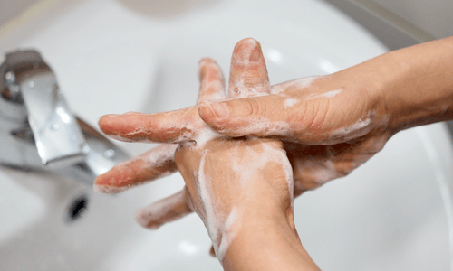 A close-up shot shows a person's hands being thoroughly washed with soap and water over a white sink. The hands are interlaced, with the fingers scrubbing the back of the opposite hand, creating a rich lather of white soap suds, demonstrating a proper handwashing technique.