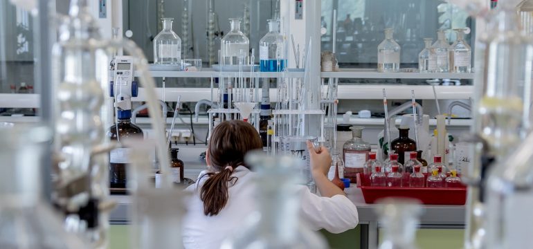 A busy chemistry laboratory scene viewed through a foreground of out-of-focus glassware. A researcher in a white lab coat with their back to the camera works at a bench, surrounded by beakers, vials, and lab equipment beneath a fume hood.