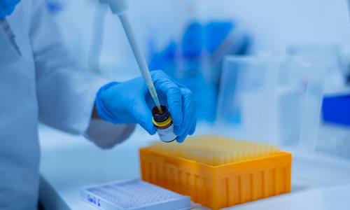 A close-up shot of a researcher wearing blue nitrile gloves and a white lab coat, carefully using a pipette to draw or dispense liquid from a small brown glass vial in a brightly lit laboratory.