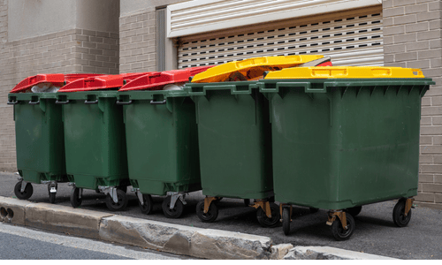 A row of large, green commercial-sized recycling or waste bins with red and yellow lids, positioned outdoors against a brick wall and a corrugated metal door.