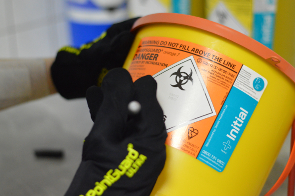Close-up of an Initial Medical sharps container with biohazard labelling, being handled by a staff member in PointGuard safety gloves to ensure correct waste segregation