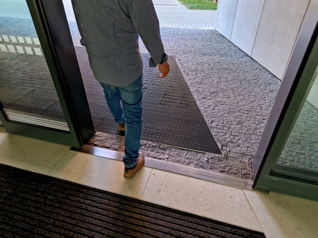 A person in jeans, brown shoes, and a striped shirt walks out through glass doors onto a textured stone walkway bordered by a building.