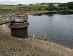 A reservoir with low water levels, showing exposed stone banks, a circular outflow structure, and metal railings. Two measuring poles stand near the water. Green fields and trees are in the background under a cloudy sky.
