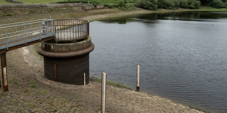 A reservoir with low water levels, showing exposed stone banks, a circular outflow structure, and metal railings. Two measuring poles stand near the water. Green fields and trees are in the background under a cloudy sky.