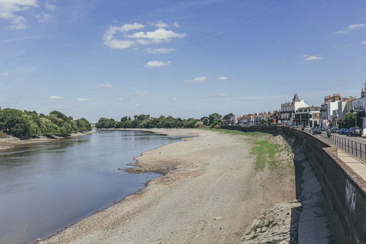 A wide, shallow river with a rocky, exposed bank runs alongside a row of houses and a street under a clear blue sky with few clouds.